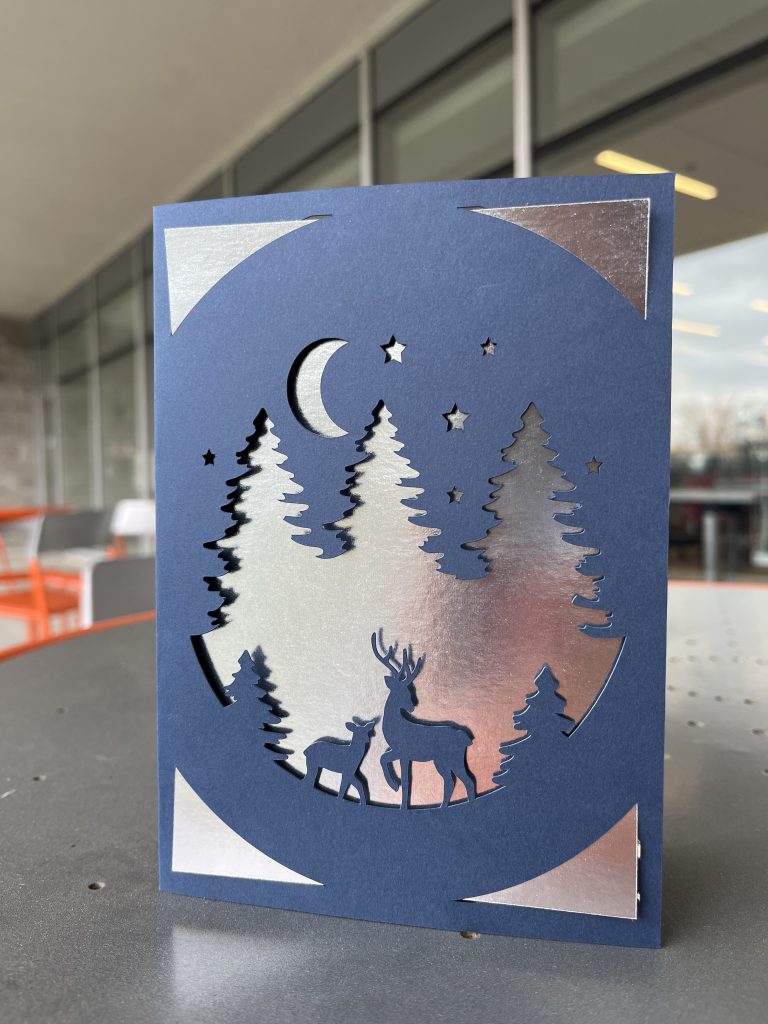 Blue and silver card featuring an adult and baby deer in front of trees with the moon in the background.
