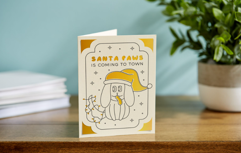 Beige and yellow card featuring a dog in a santa hat and text that reads "Santa Paws is coming to town"